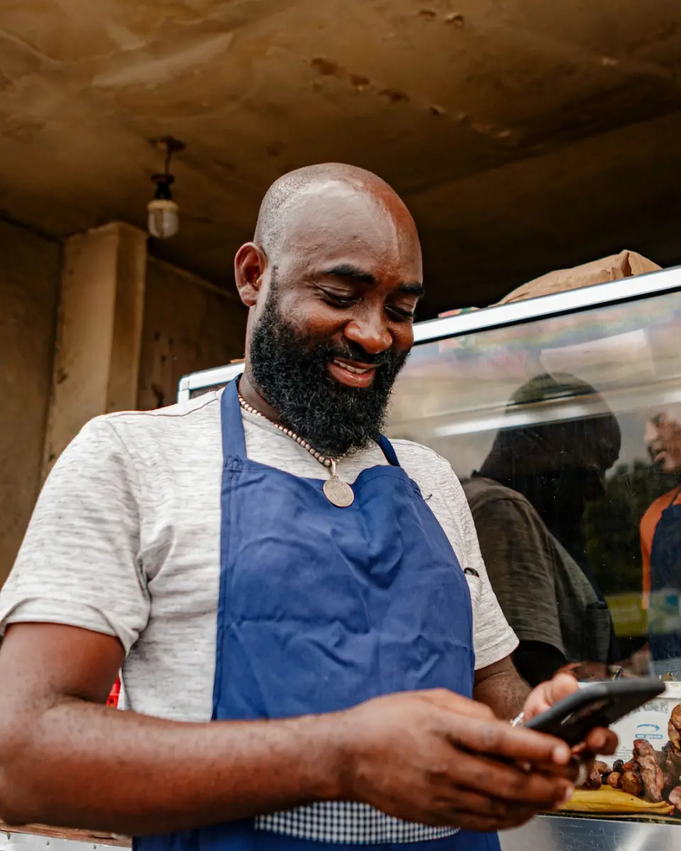 A food stand owner in a blue apron smiles while checking a phone